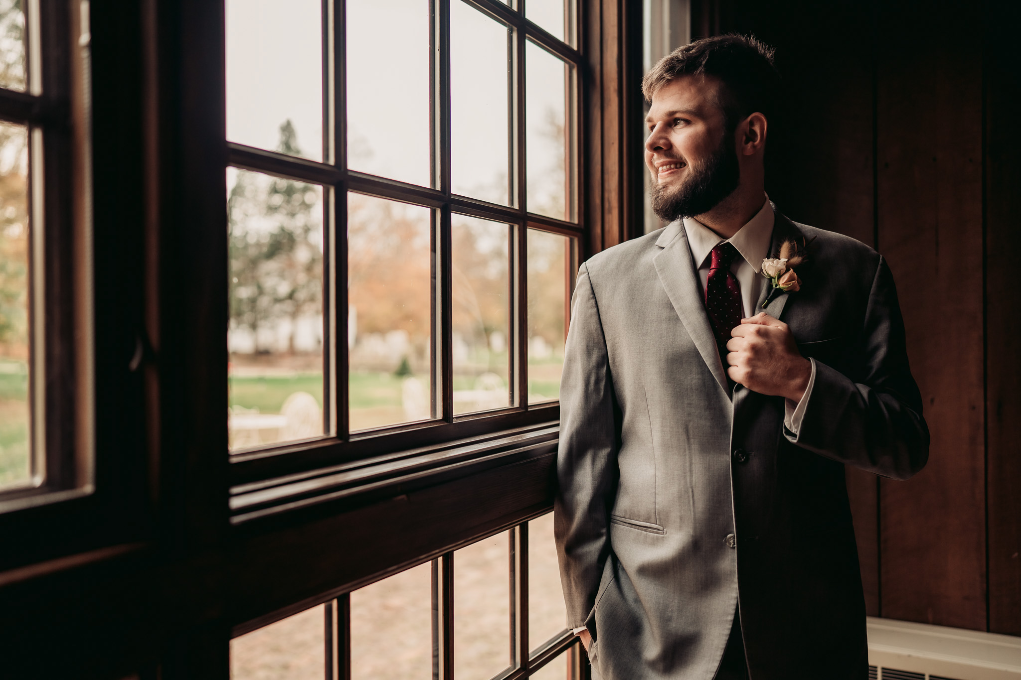 Color photograph of a groom in a gray suit posing before a wedding ceremony on a cloudy fall day by a window holding his lapel and smiling inside of a building at Waterloo Village in Byram New Jersey 