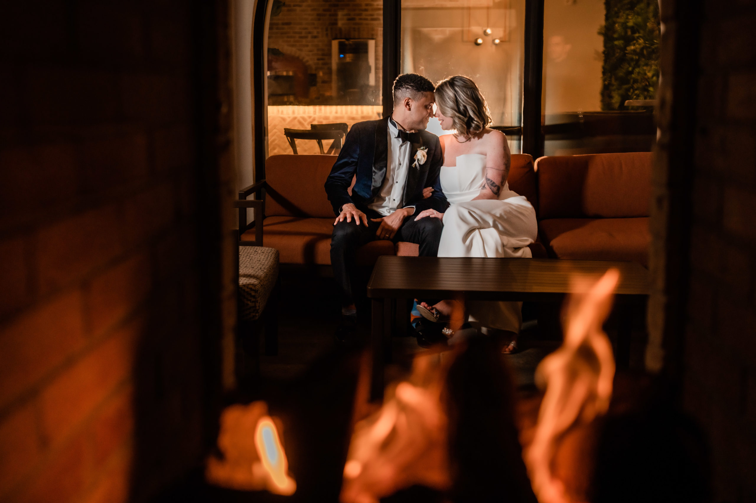 A photographic portrait of a couple sitting on an orange couch outside on a patio embracing and putting their heads together at night time by a fire place on the patio during their wedding in Andover New Jersey.