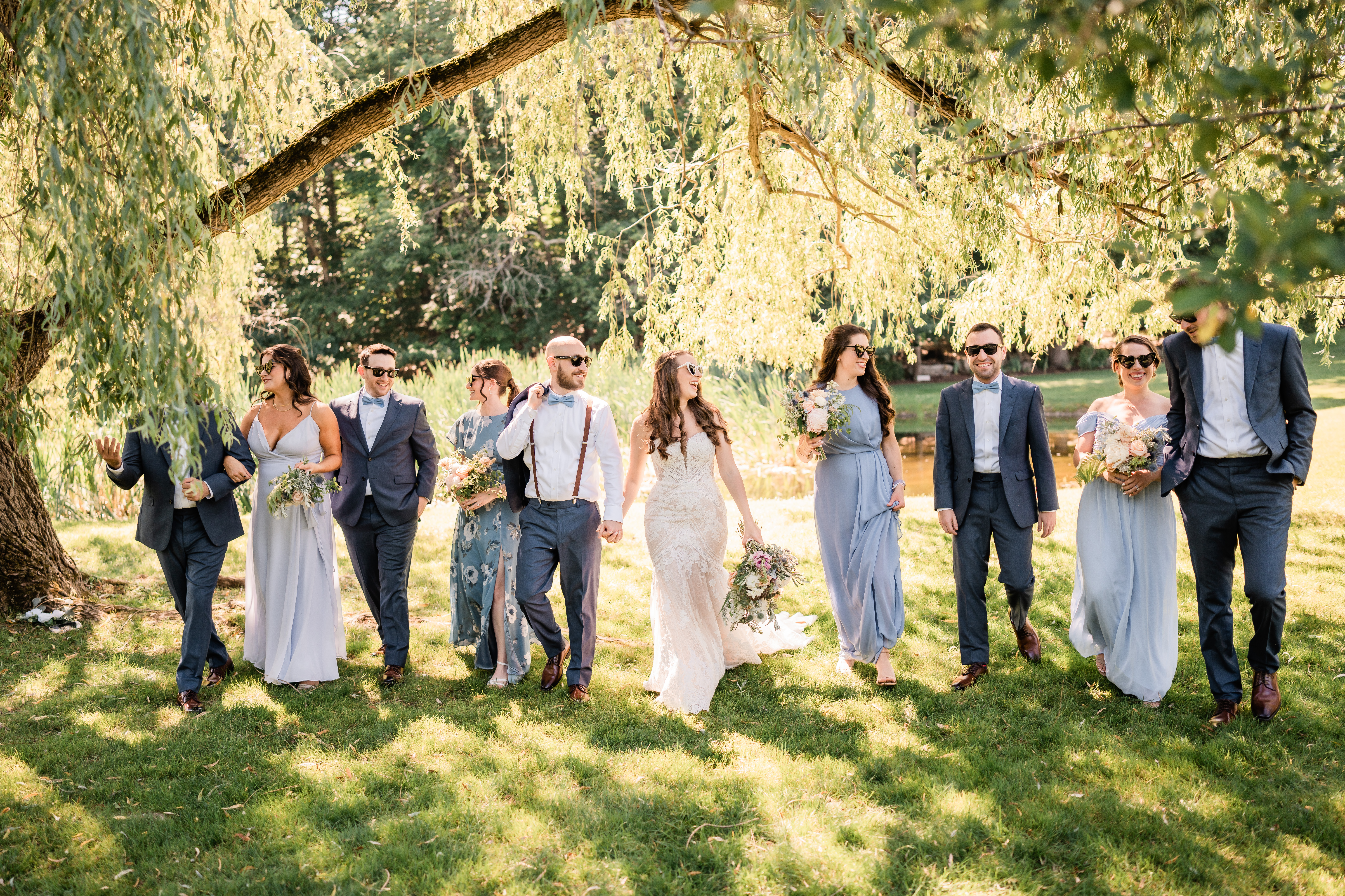 Perona Farms Wedding, Perona Farms Wedding Photographer, NJ Weddings, New Jersey Wedding Photographer, Perona Farms Refinery Wedding Photographer, NJ Wedding Photographer, Perona Farms Reserve Wedding Photographer, New Jersey Wedding Venues, Perona Farms Barn Wedding Photography, Wedding Inspiration, Wedding Planning, Unique Wedding Photos, Wedding Photo Ideas, group photograph of bride and groom wearing sunglasses and walking + laughing with their wedding party under a Willow tree at Perona Farms