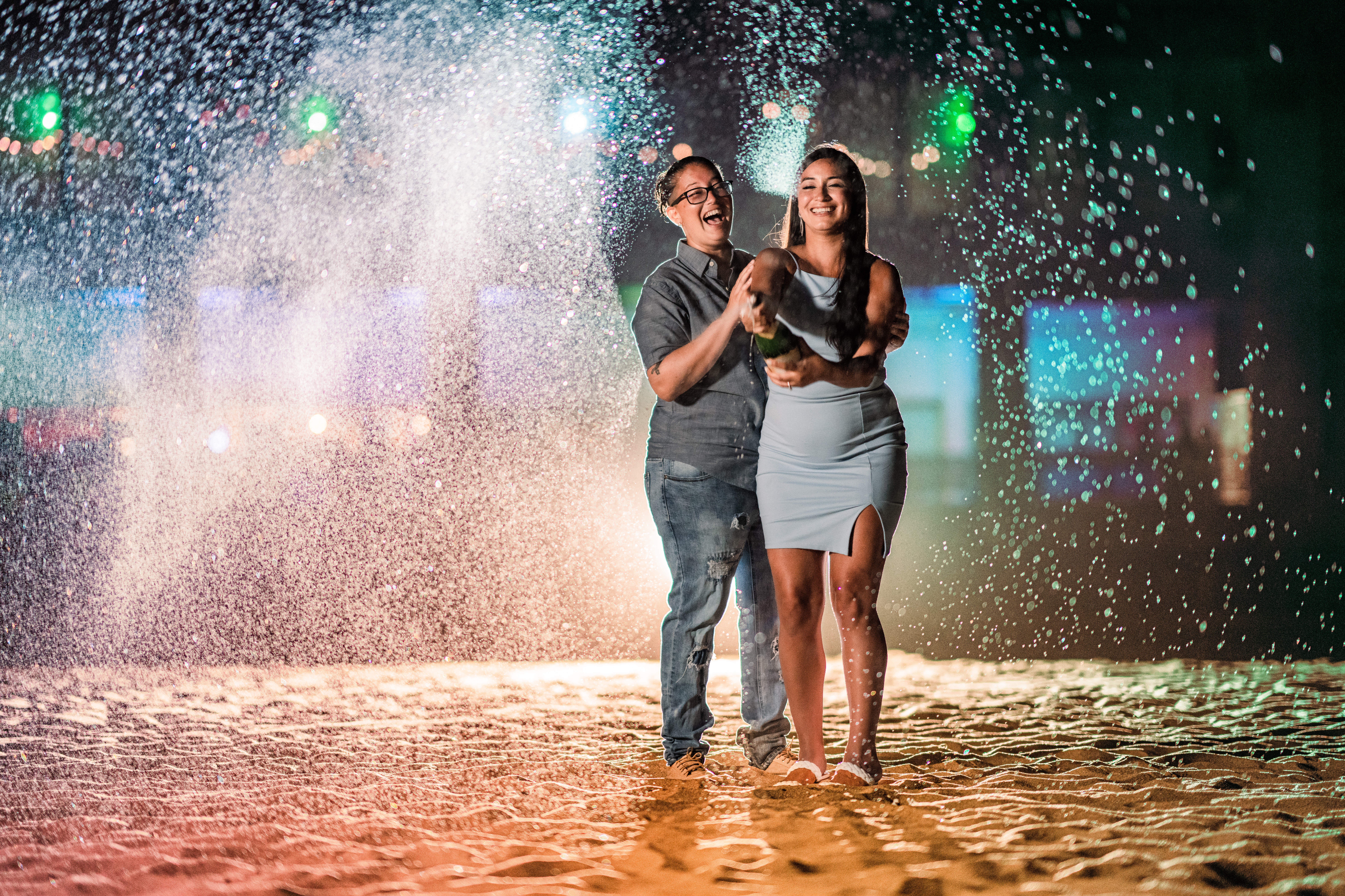 Asbury Park Engagement, Asbury Park Proposal, Asbury Park Photographer, Asbury Park Proposal Photographer, LGBTQ couple smiling while popping champagne on the beach while being backlit with a flash 
