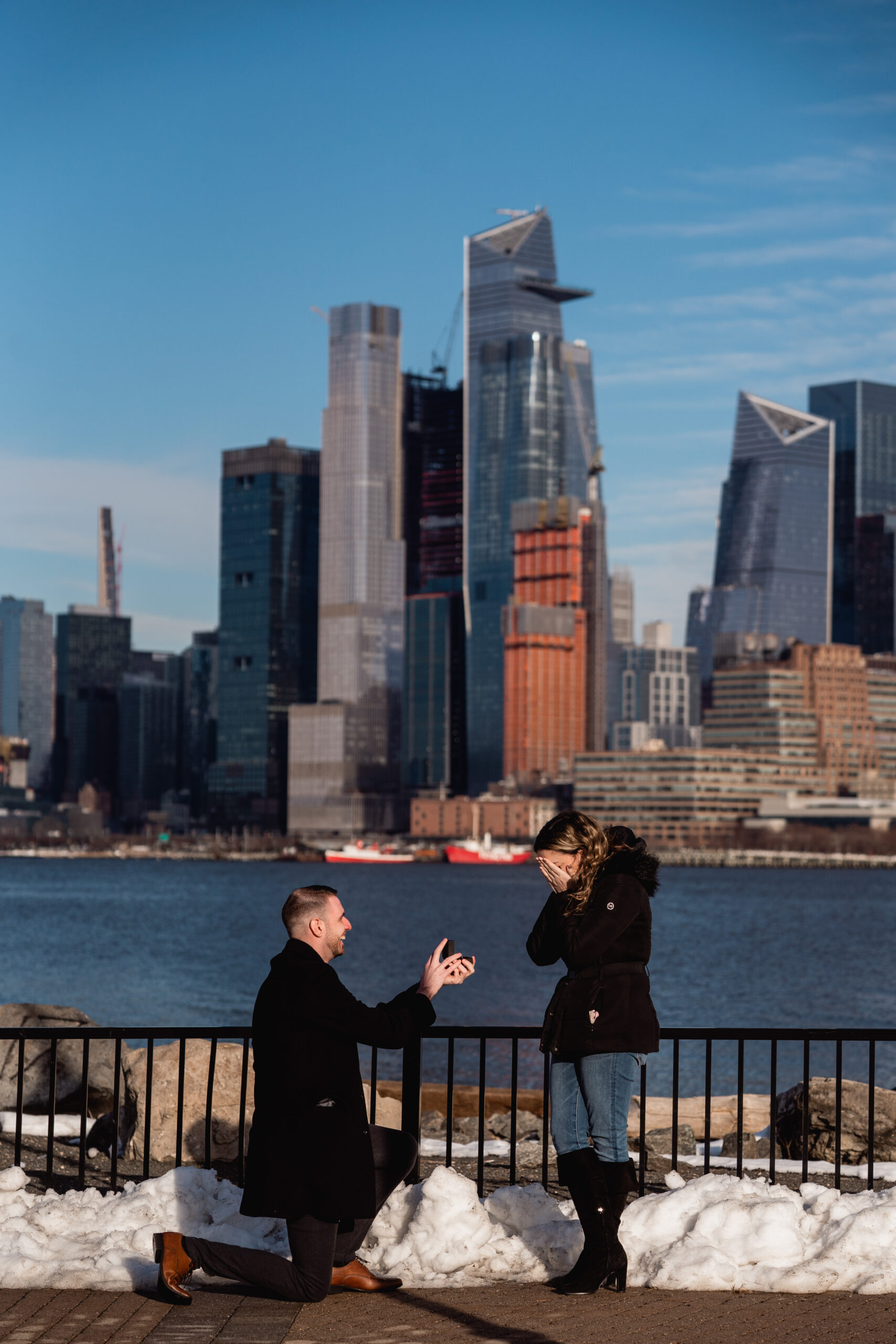 Hoboken New Jersey Proposal, Hoboken Proposal, Hoboken Proposal Photographer, Hoboken Photographer, New Jersey Photographer, NJ Engagement Photographer, NJ Winter Proposal, man kneeling down to propose to his fiancé as she covers her face in shock with the NYC skyline in the background and snow around them 
