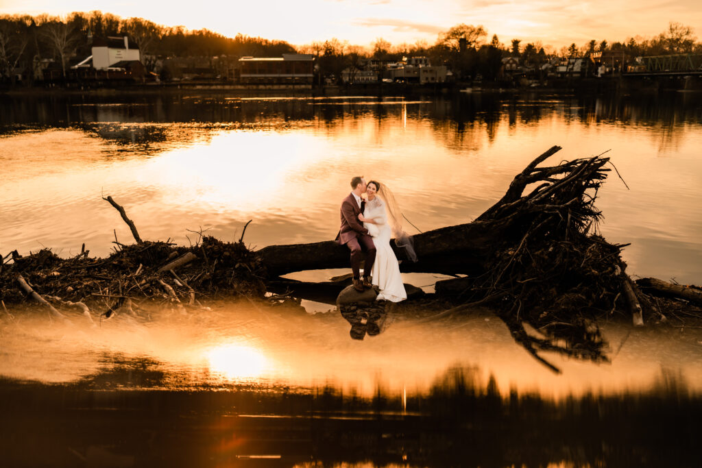 Lambertville Station Inn Wedding Photographer, NJ Weddings, New Jersey Wedding Photographer, NJ Wedding Photographer, New Jersey Wedding Venues, Wedding Photography, Wedding Inspiration, Wedding Planning, Wedding Ideas, Unique Wedding Photos, Wedding Photo Ideas, Bride and Groom hugging and kissing while sitting on a fallen tree along the Delaware River.