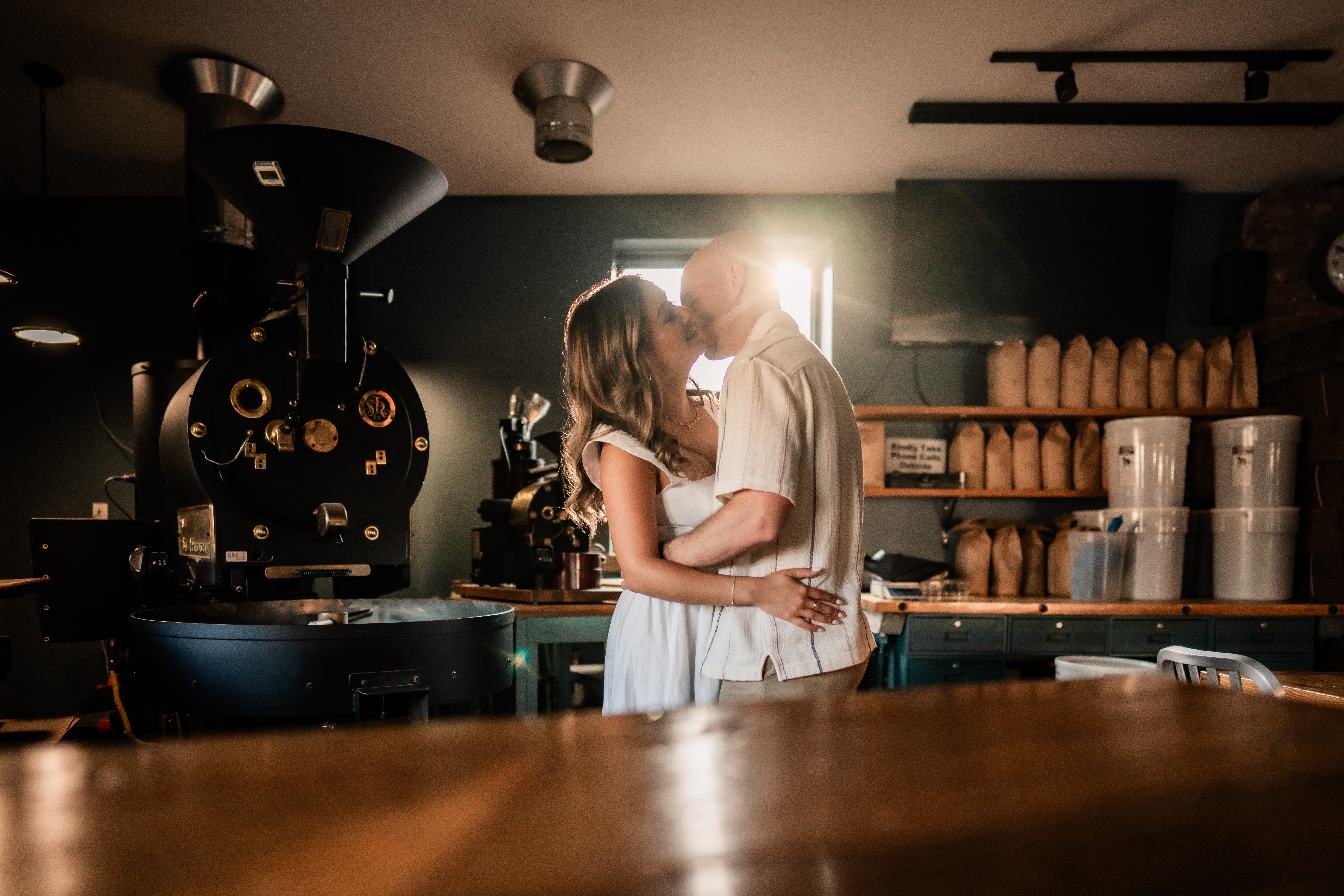 New Jersey engagement photography at a Belmar coffee shop, Turnstile Coffee Roasters, with an engaged couple kissing beside a large coffee roaster in warm window light.