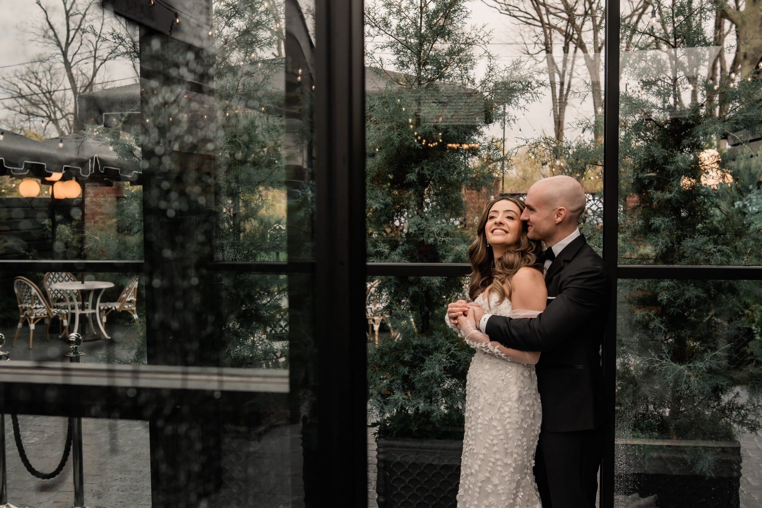 Rainy-day Park Loft wedding photography in New Jersey, with the bride laughing as the groom embraces her beside the venue’s glass windows and greenery.
