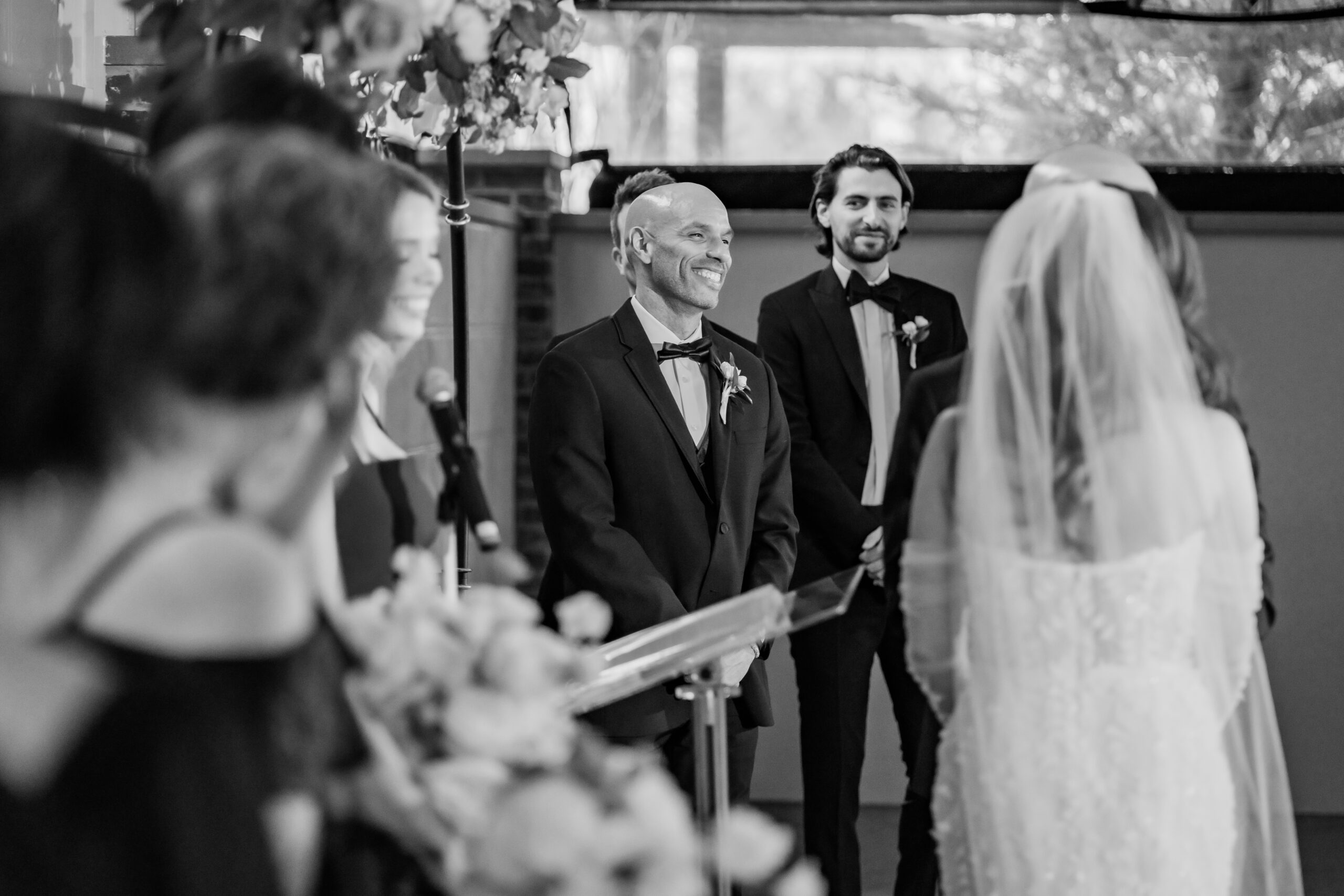 Black-and-white Park Loft wedding photo of the best man smiling at the couple during their ceremony in the Belvedere room.
